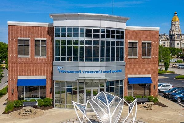 Aerial shot of the two-story Indiana State University Foundation building. Red brick is visible on the sides with a white and silver center façade featuring glass windows and doors. The Foundation’s name in blue letters appears between the first and second floors. There is a large Plexiglass sculpture of a Sycamore leaf in front of the building. The Vigo County Courthouse is visible in the distance on the left.